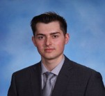 man with dark hair wearing dark suit in front of blue background