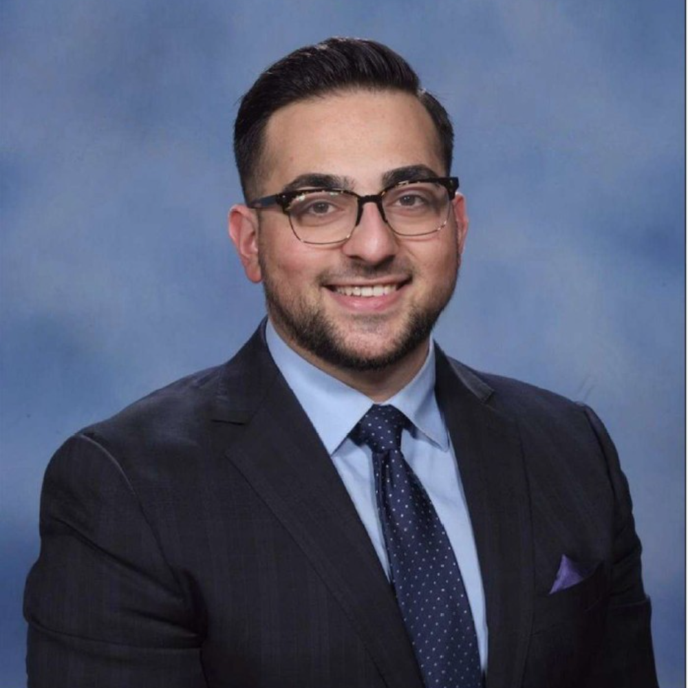 man with beard wearing glasses wearing a dark suit, blue shirt and tie