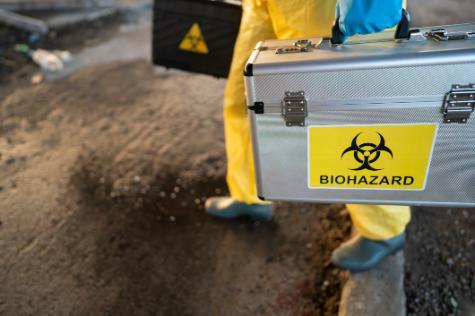 A person in PPE carries a metal case with the biohazard symbol in black and yellow on it.