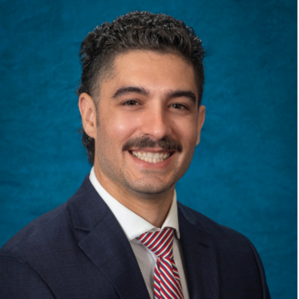 man with dark hair and mustache wearing dark suit and tie in front of blue background