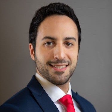 man with dark hair and beard wearing dark suit with red tie