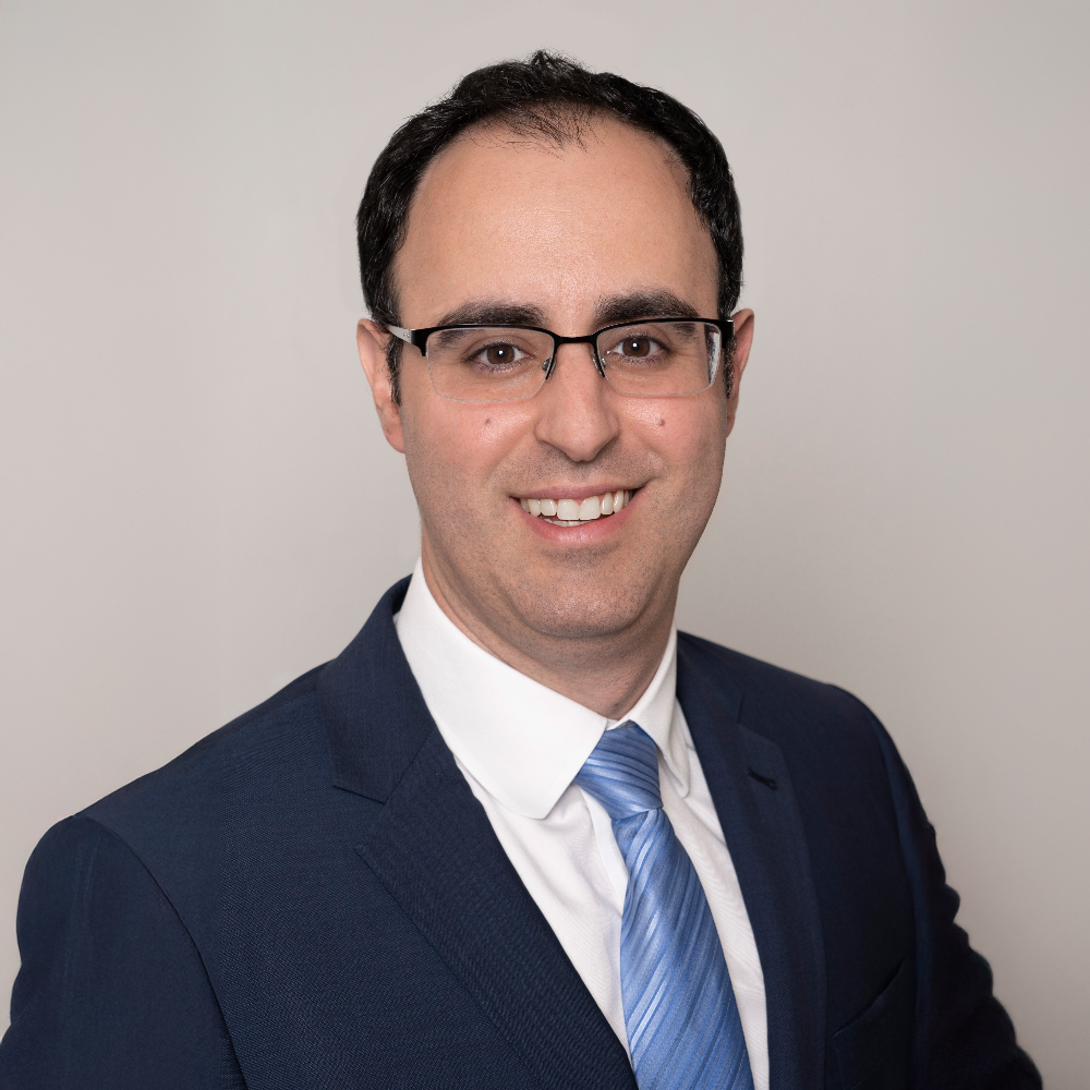 Man with dark hair wearing glasses, dark blue suit with white shirt and blue tie