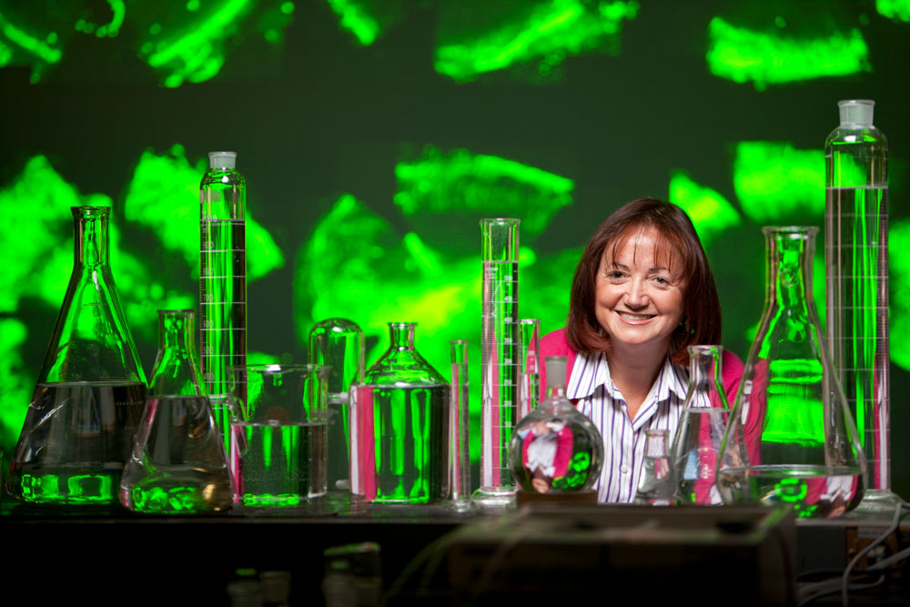 Portrait of Michal Schwartzman Ph.D., smiling, wearing a striped blouse, sitting in front of test tubes and beakers.