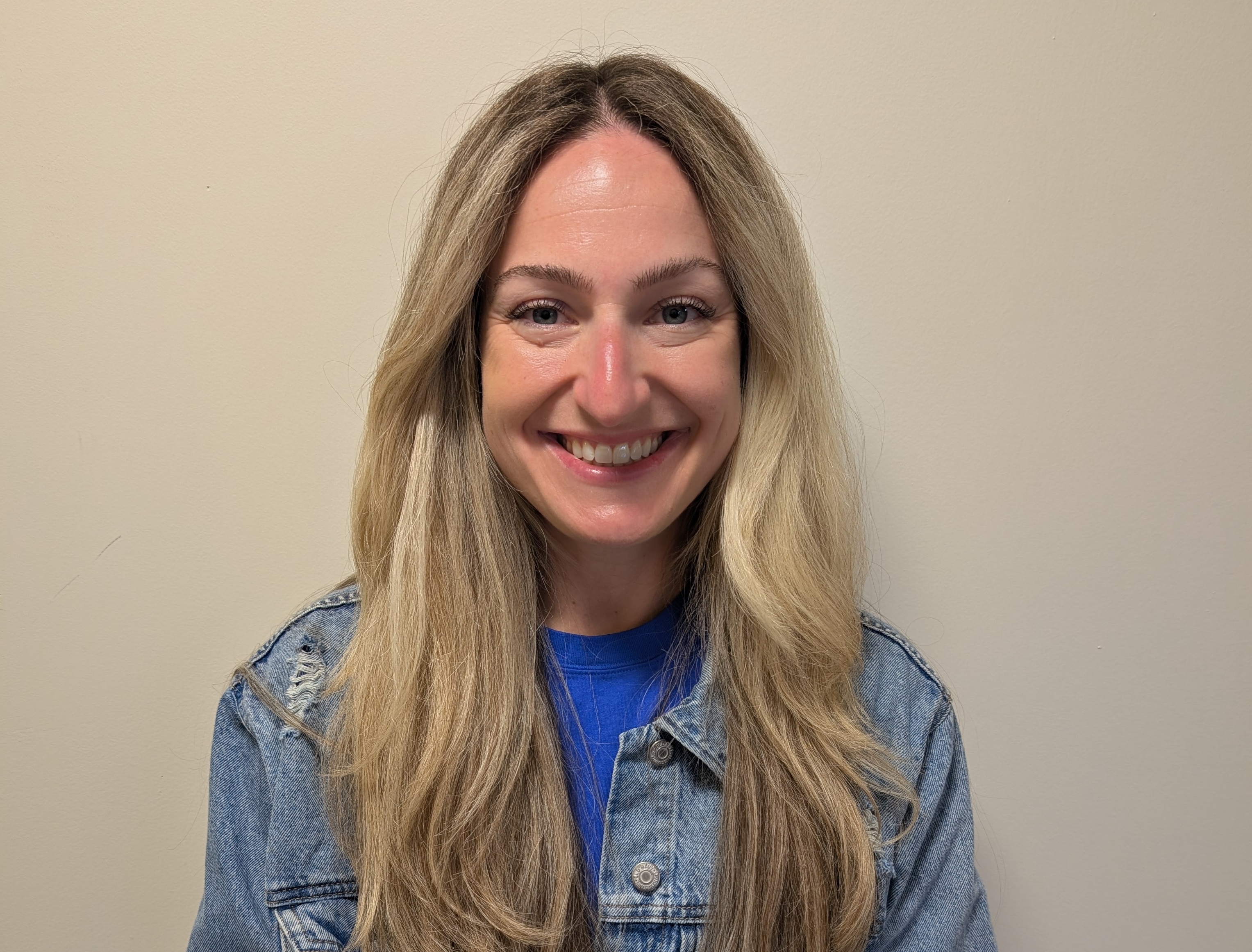 A woman with long blond hair wearing blue jean jacket smiling in front of beige background