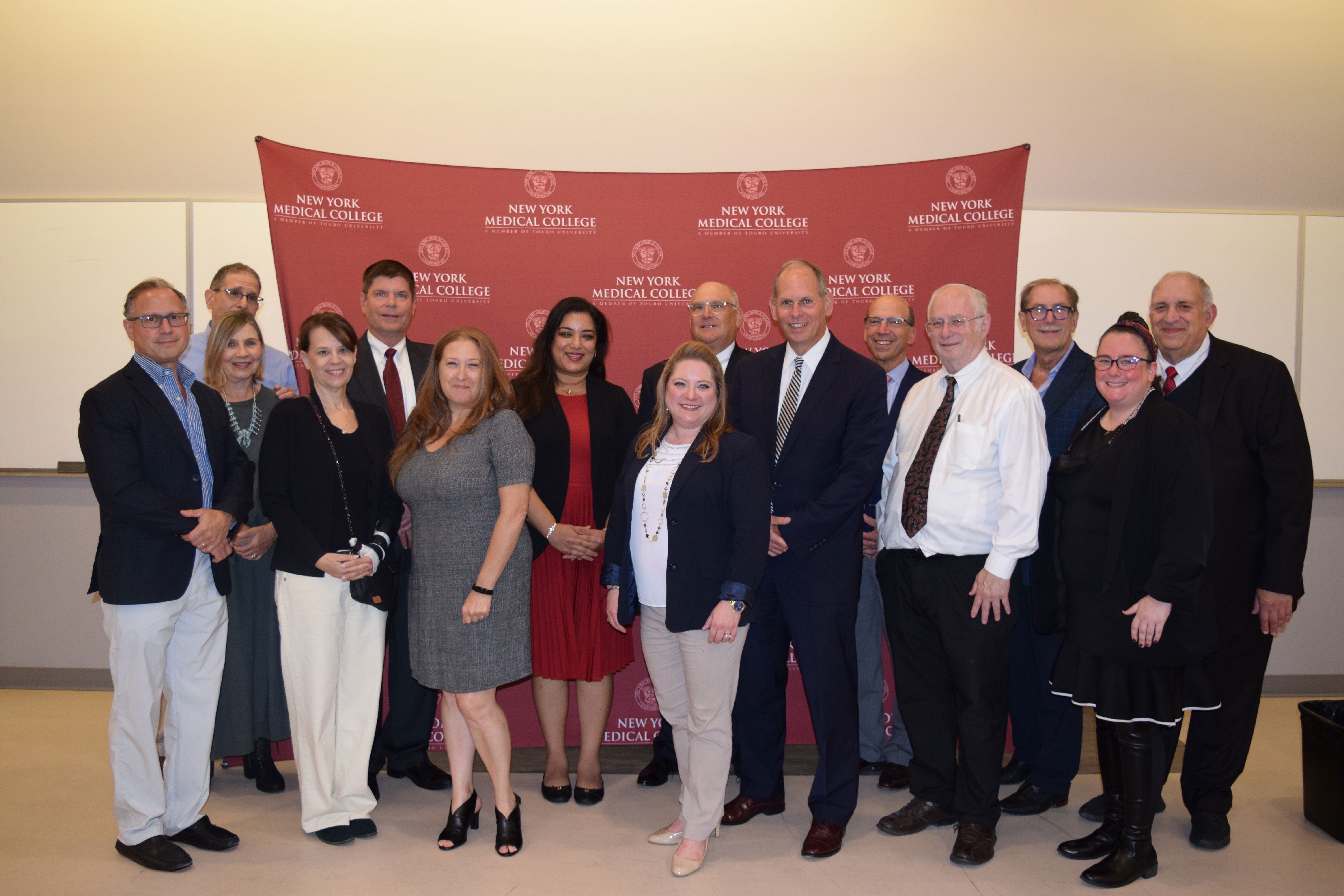 Sixteen men and women wearing suits and dresses smiling in front of a maroon New York Medical College backdrop.