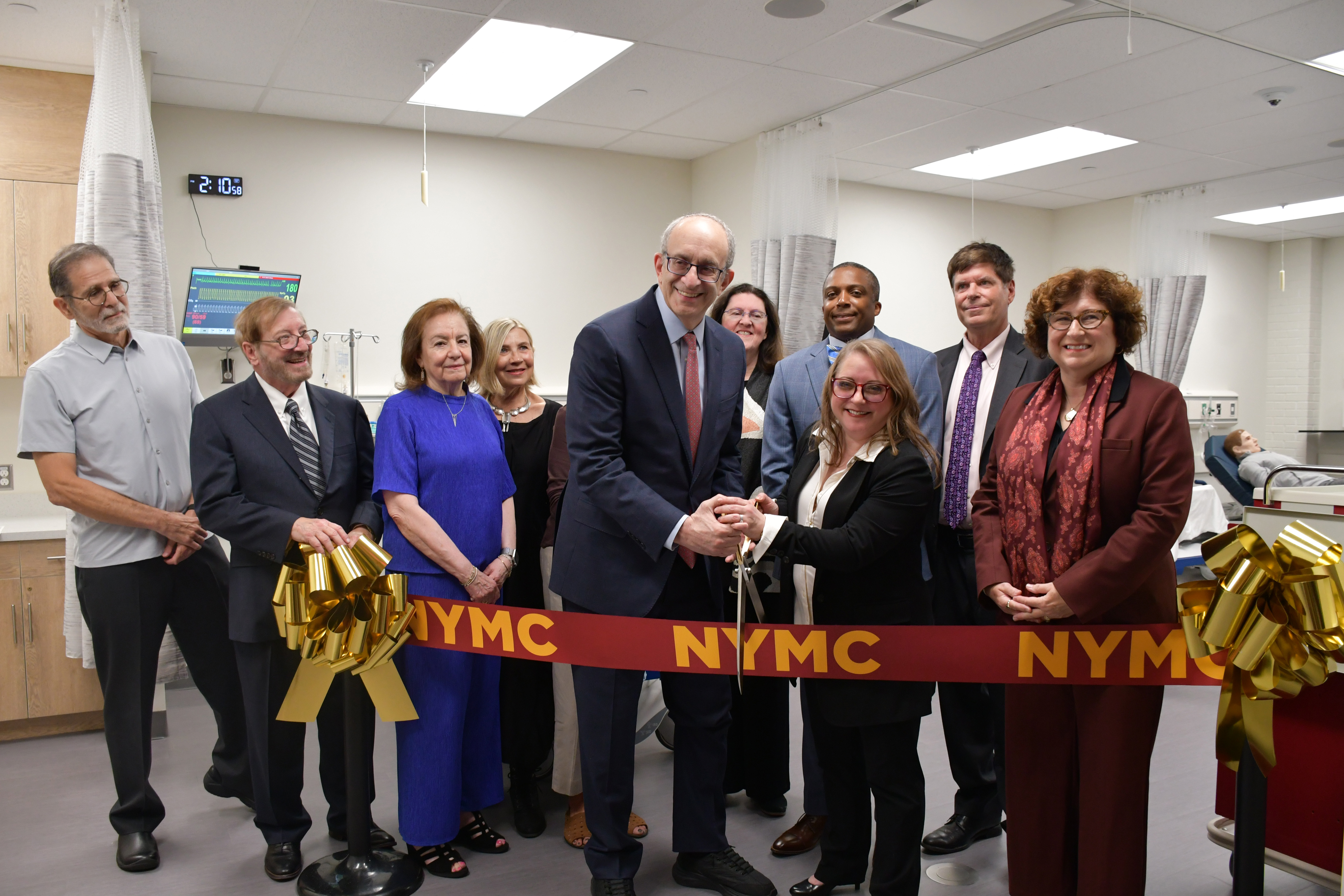 Men and women in business attire in a hospital-like setting with manikins in the background getting ready to cut a ribbon.