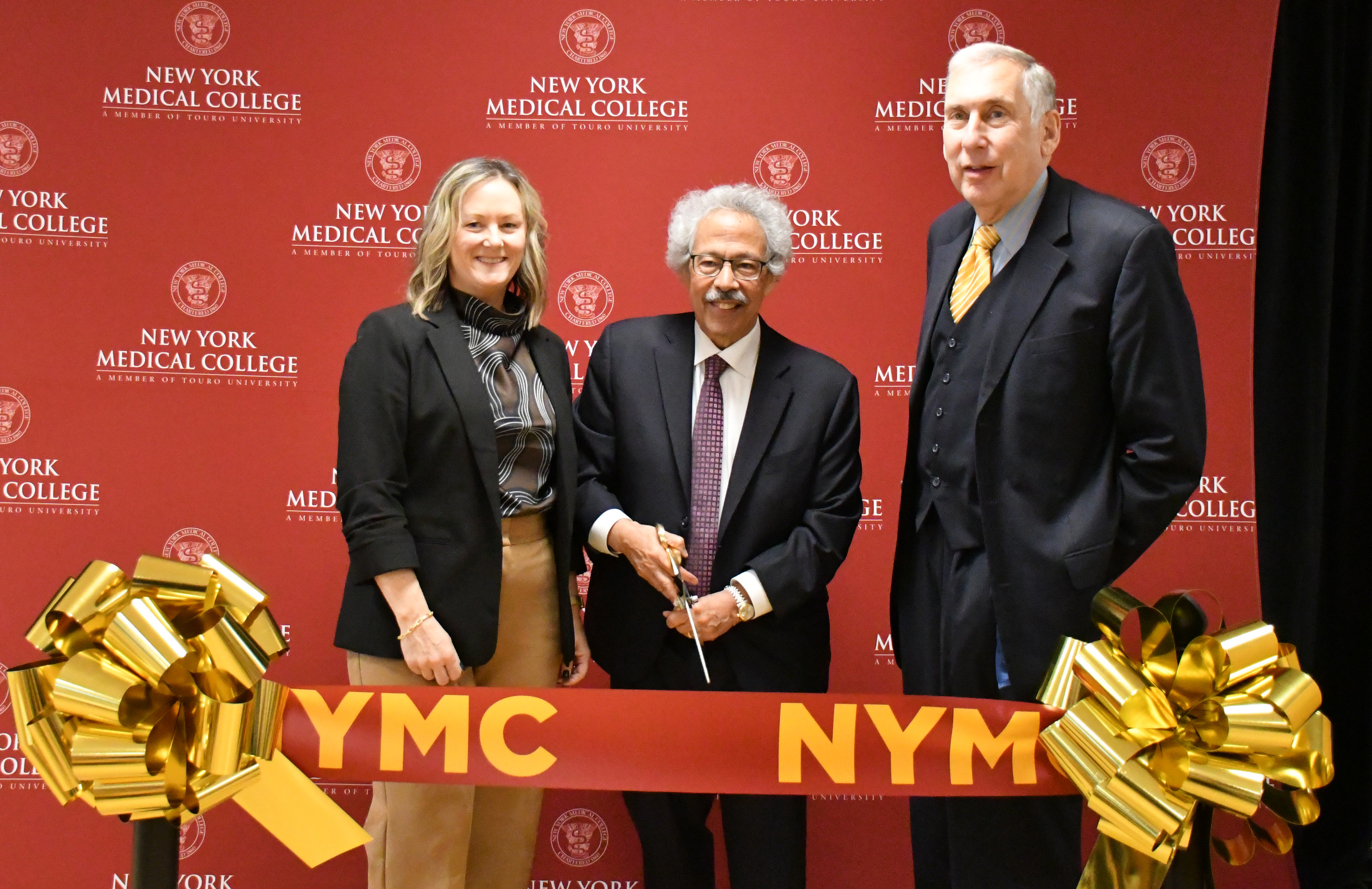 A woman and two men standing in front of a step and repeat. The man in the middle holds scissors to cut celebratory ribbon.
