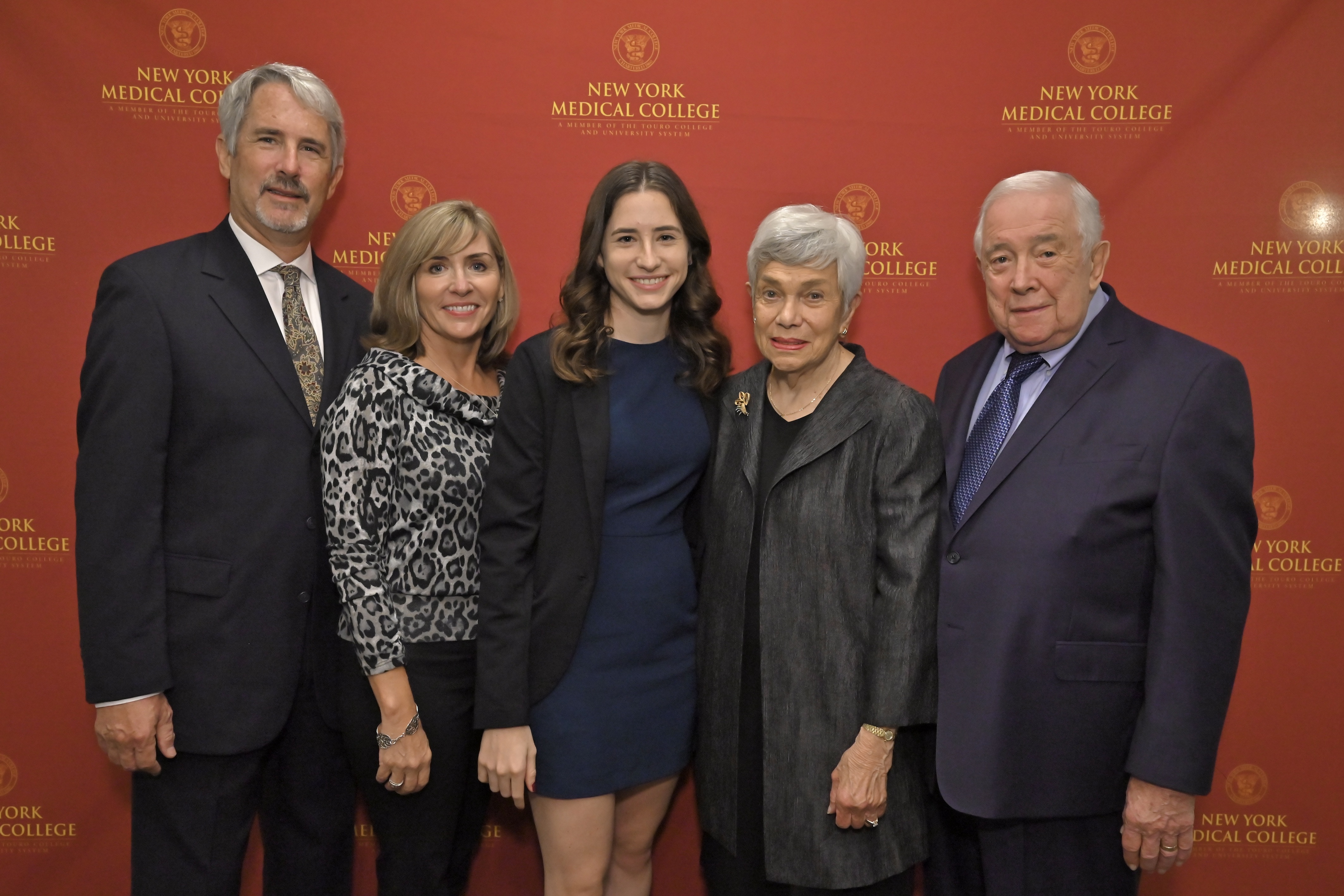 A father, mother, adult daughter and grandparents smiling in formal attire in front of a step and repeat.