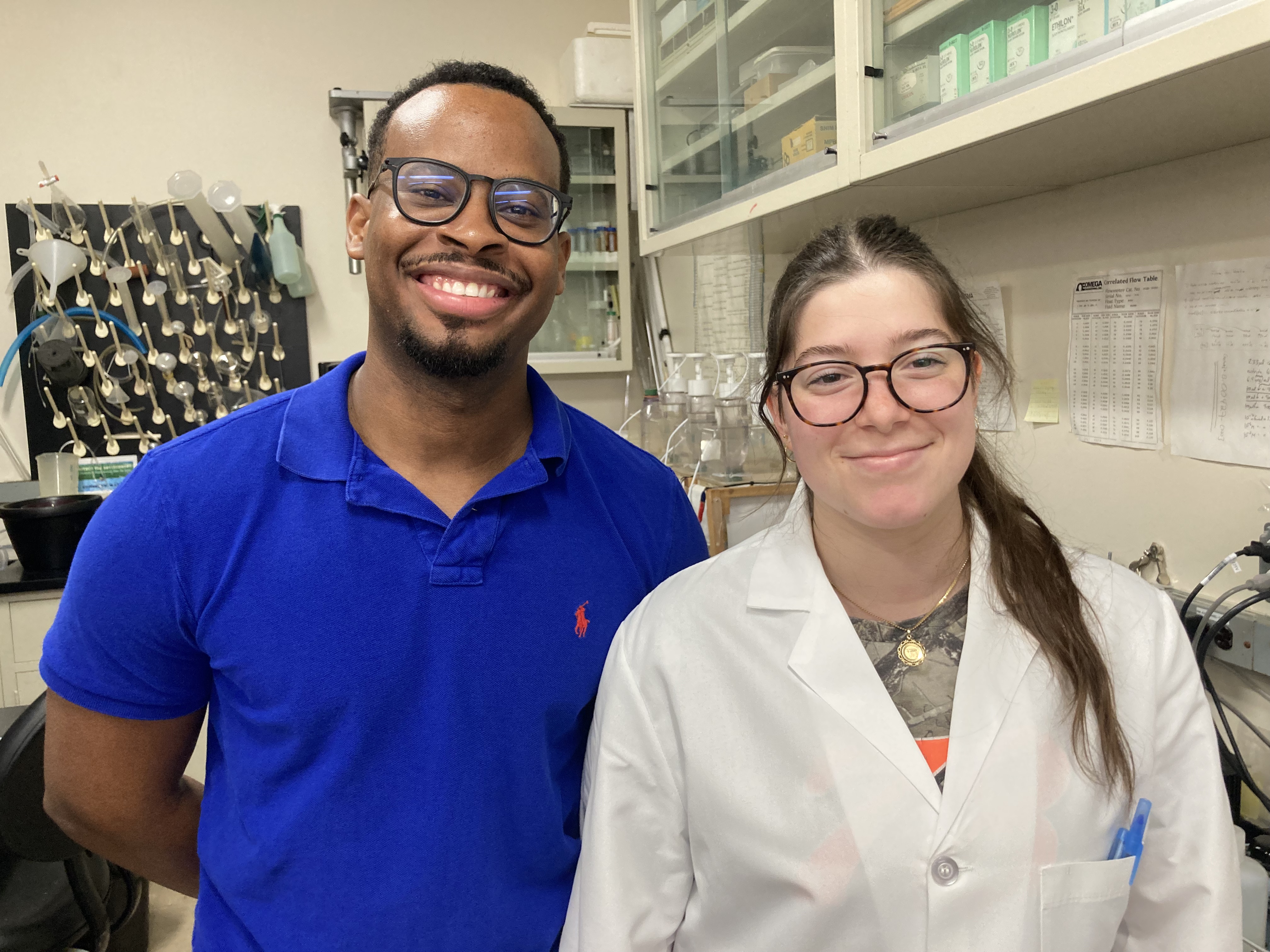 A male student with glasses and a polo shirt in a research lab with a female student wearing glasses and a white lab coat