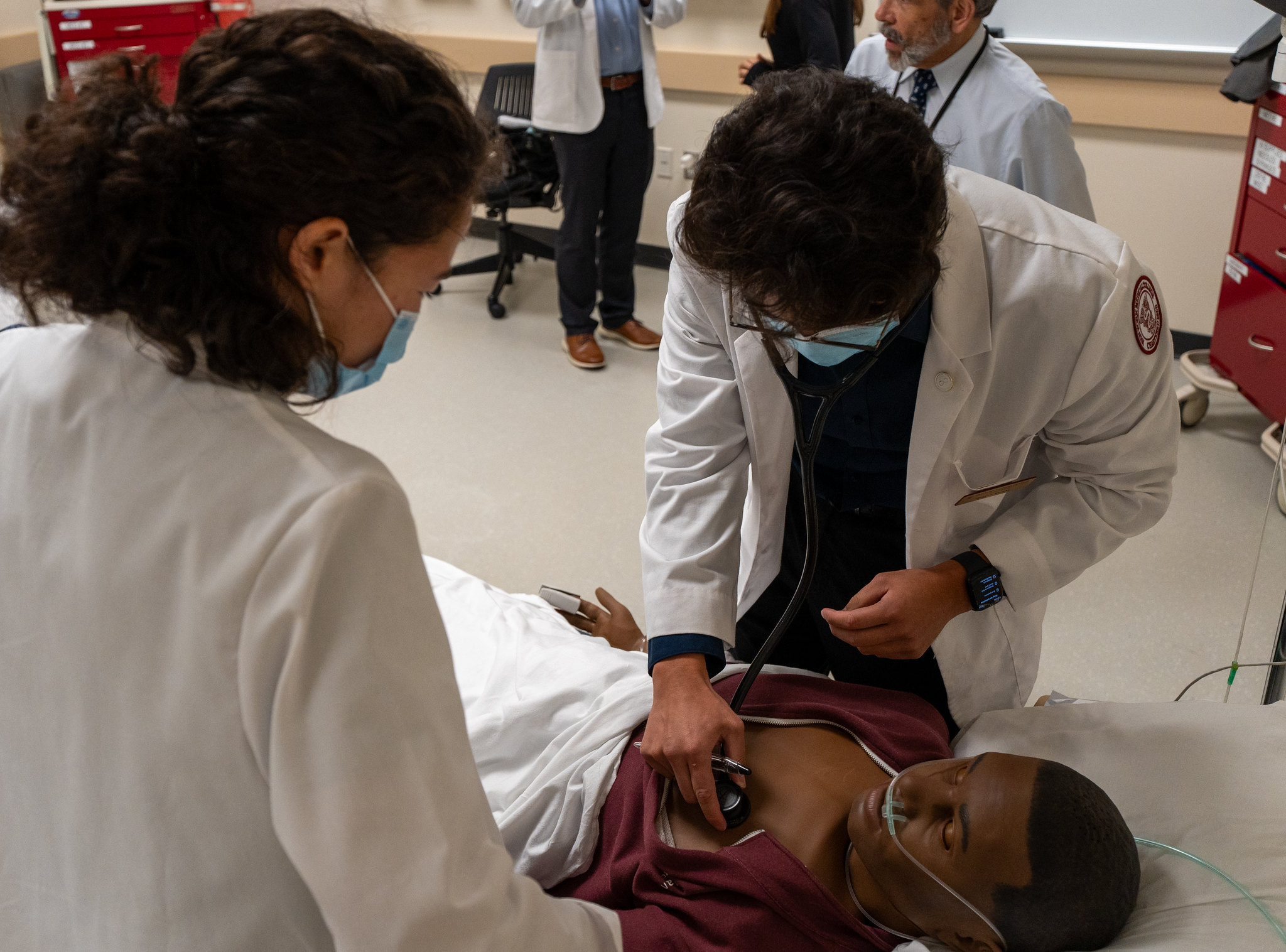 Two medical students wearing white lab coats practicing on a dark skin manikin with a stethoscope.