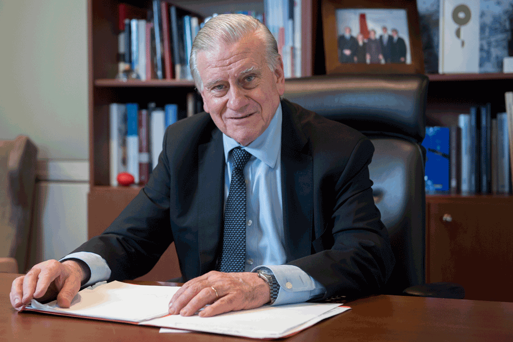 Dr. Valentin Fuster is wearing a suit, sitting at his desk with papers and a bookshelf behind him.