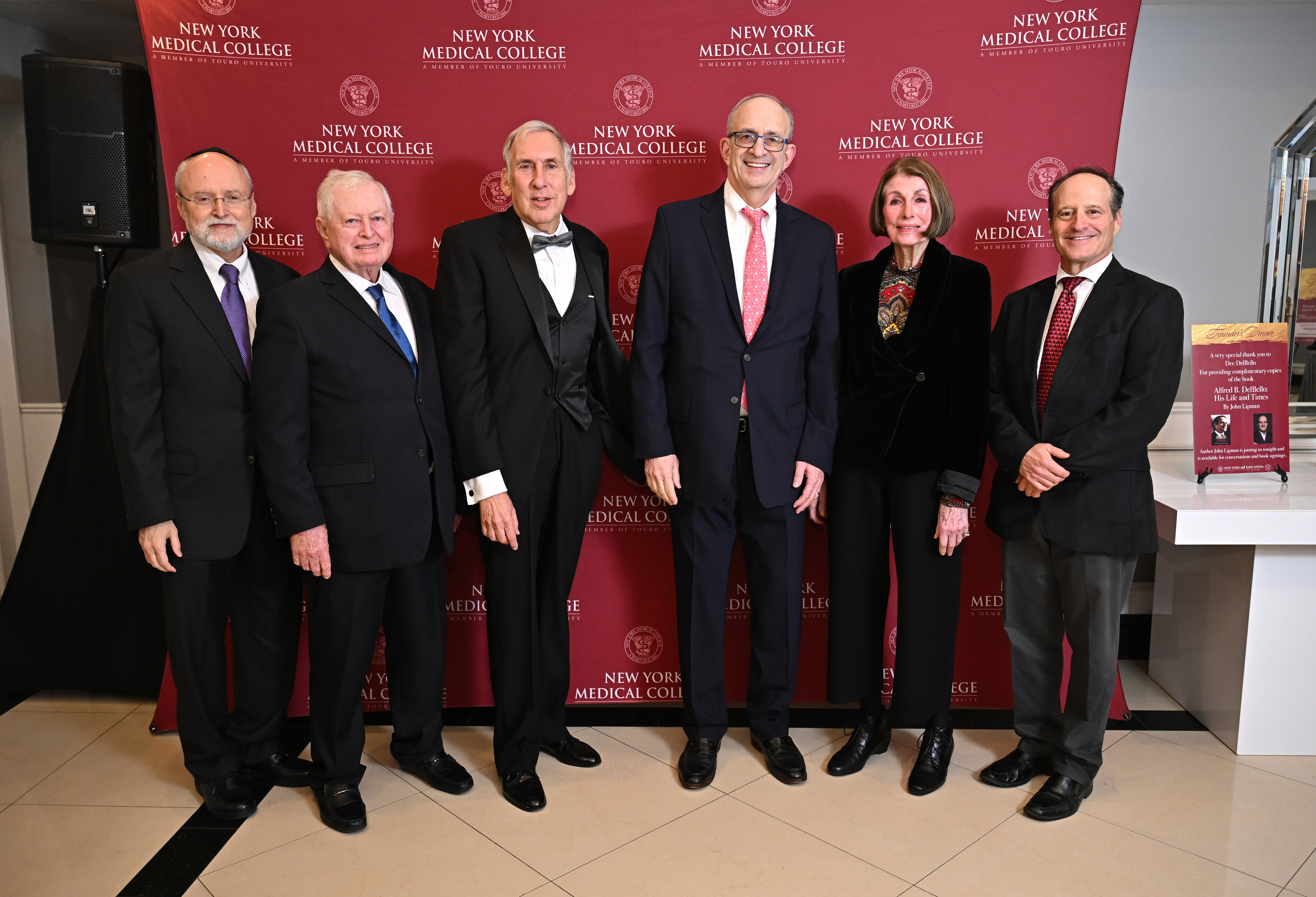 Six attendees in formal evening attire stand in front of a red New York Medical College step-and-repeat backdrop at a reception event. The group poses shoulder to shoulder for a photo, with NYMC logos displayed repeatedly across the background.