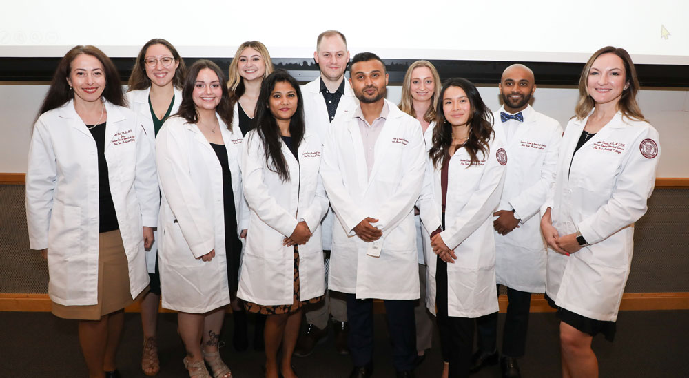 A dozen students and faculty wearing white lab coats smiling in front of a white projector.