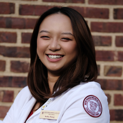 Woman smiling facing forward wearing a white coat in front of a brick wall