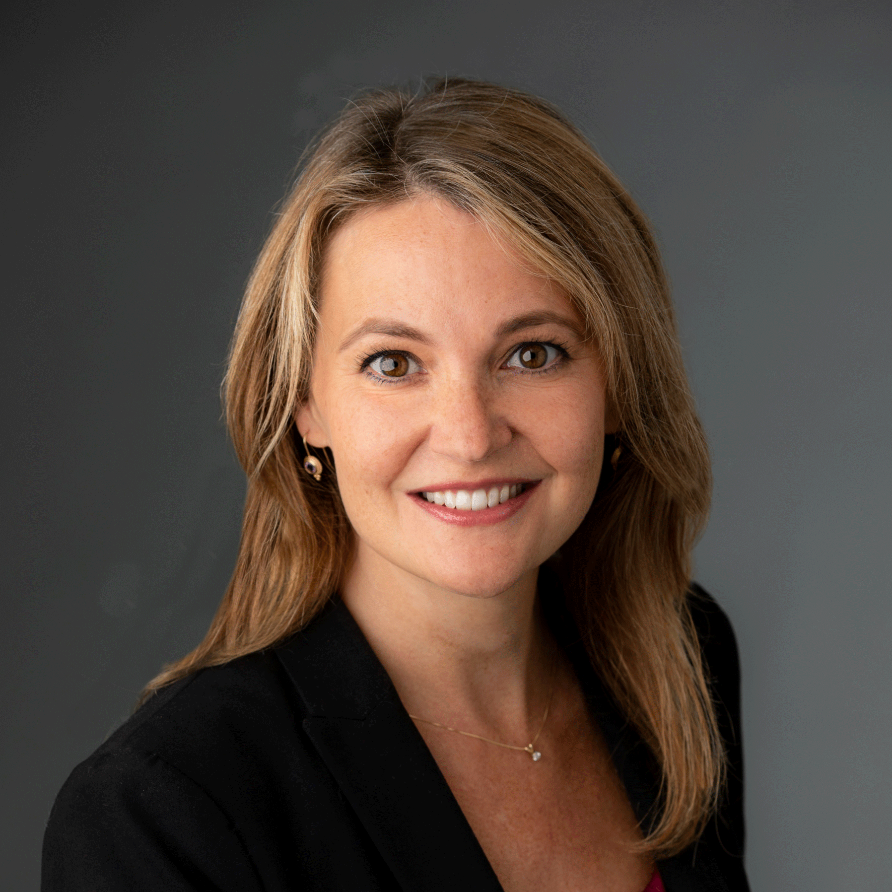 Woman with blond hair smiling with a black suit jacket and fuchsia shirt in front of a gray background