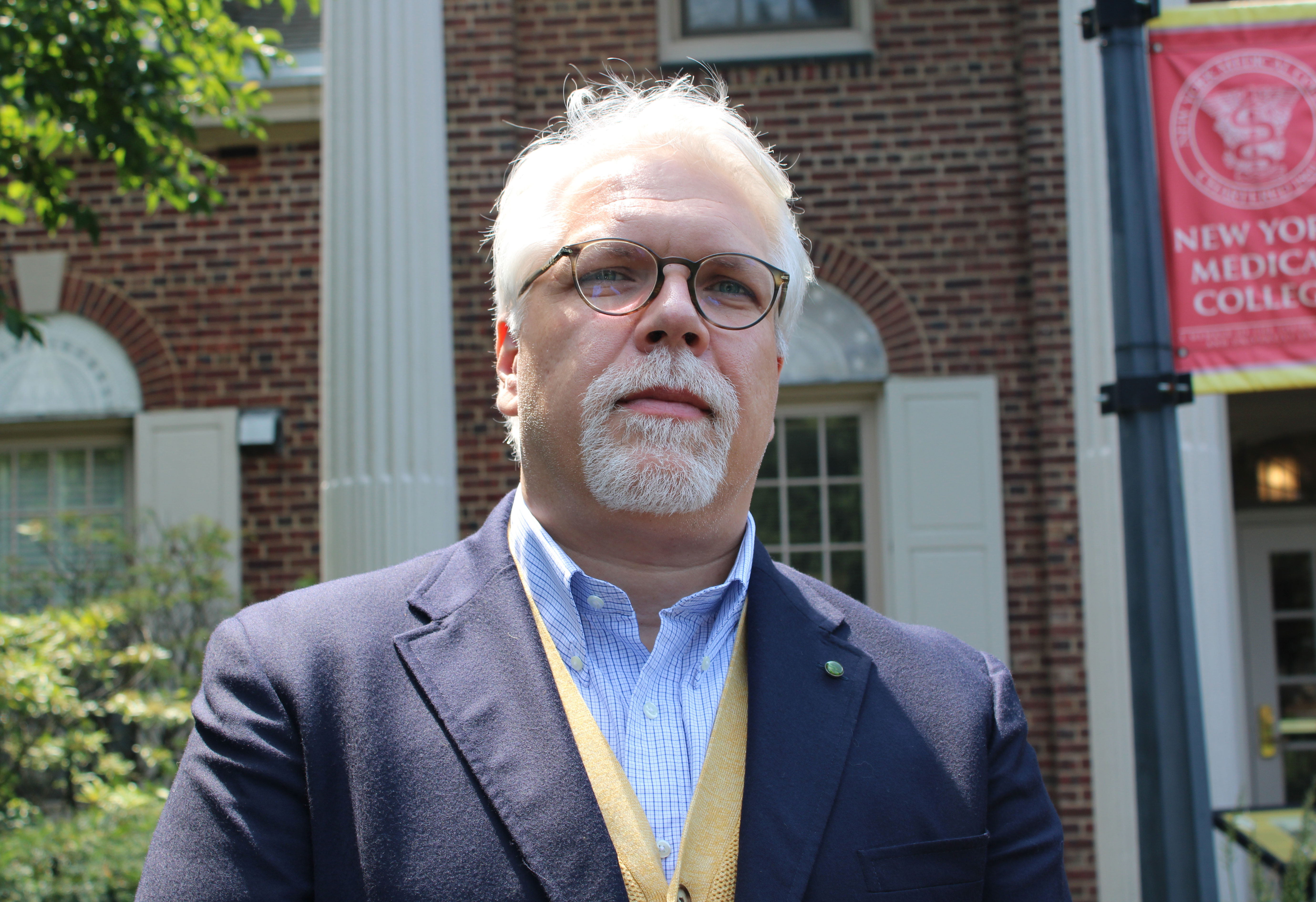 Man with white hair and beard wearing a navy blue suit outside on a sunny day in front of a building