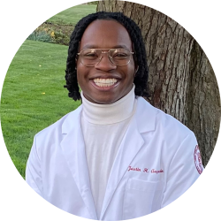 Male medical student with shoulder length twists and glasses wearing a turtleneck and white lab coat smiling outside in front of a tree
