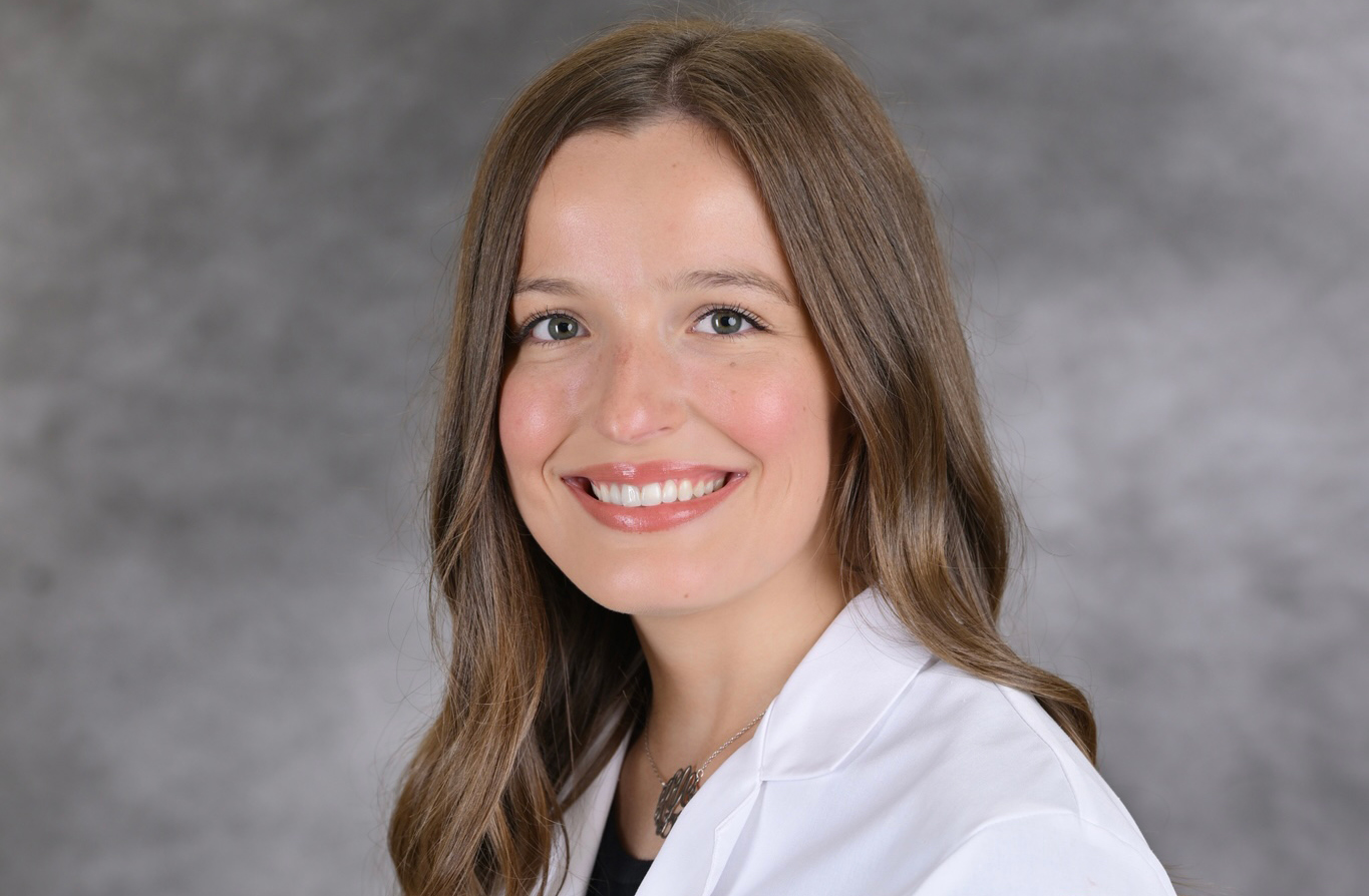 A female student with long hair smiling in a white coat in front of gray backdrop