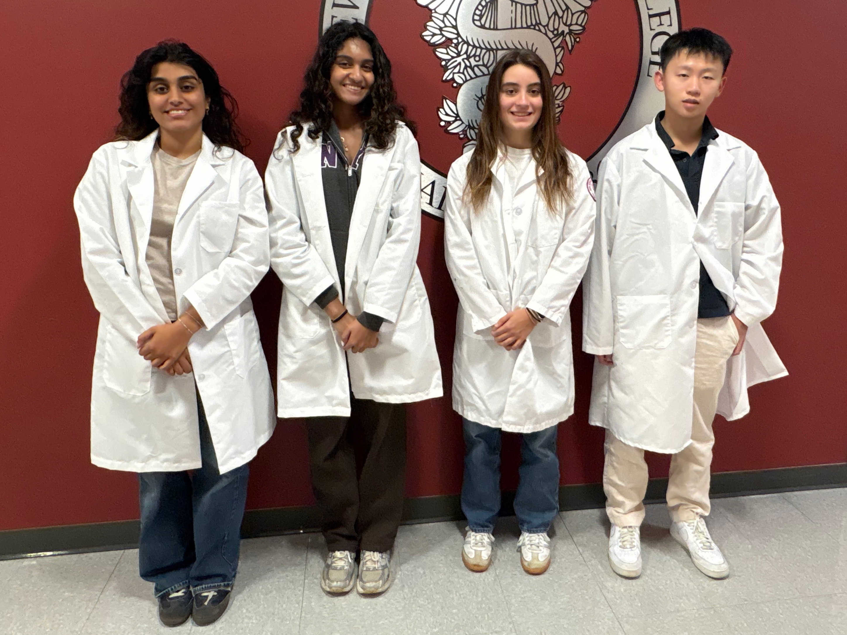 Three women and one man student wearing white coats smiling in front of New York Medical College emblem