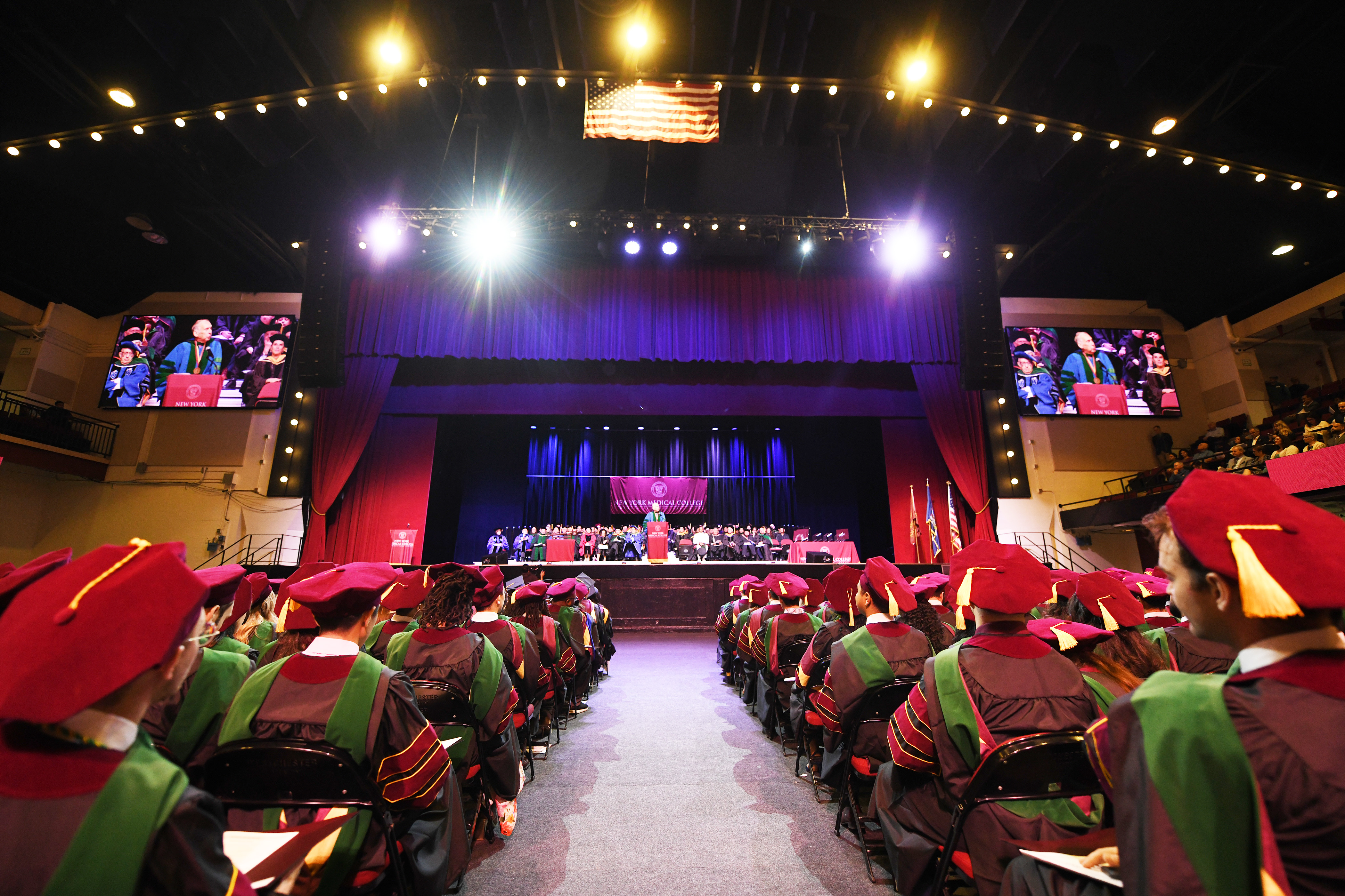 Westchester County Center with doctoral and master\'s students sitting in their cap and gowns facing a stage with faculty in regalia and a man at the podium.