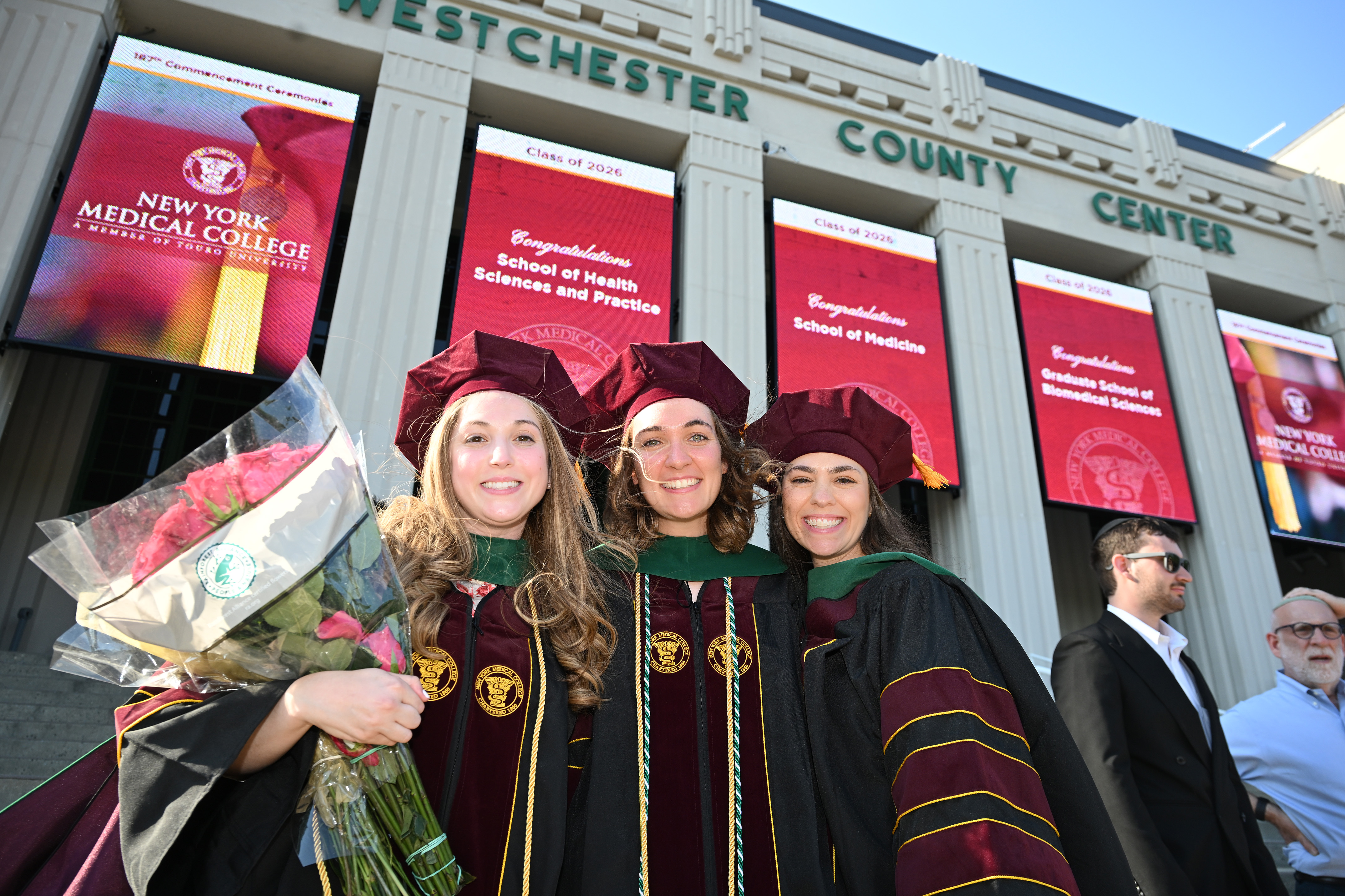 Three women in regalia smiling together in front of Westchester County Center