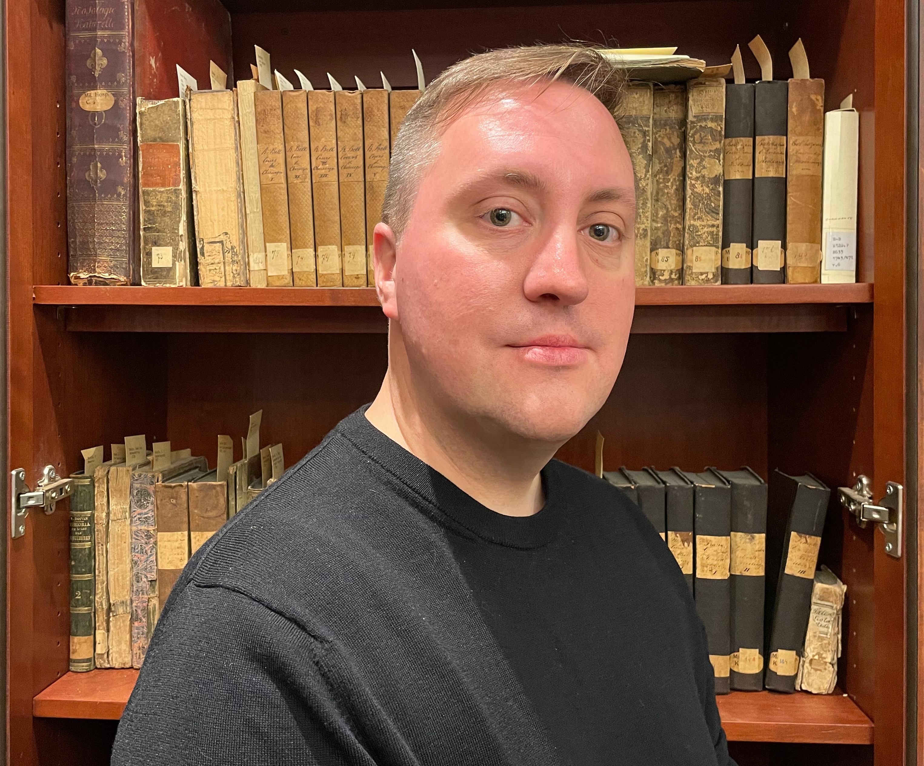 Man wearing a dark shirt softly smiling in front of books.