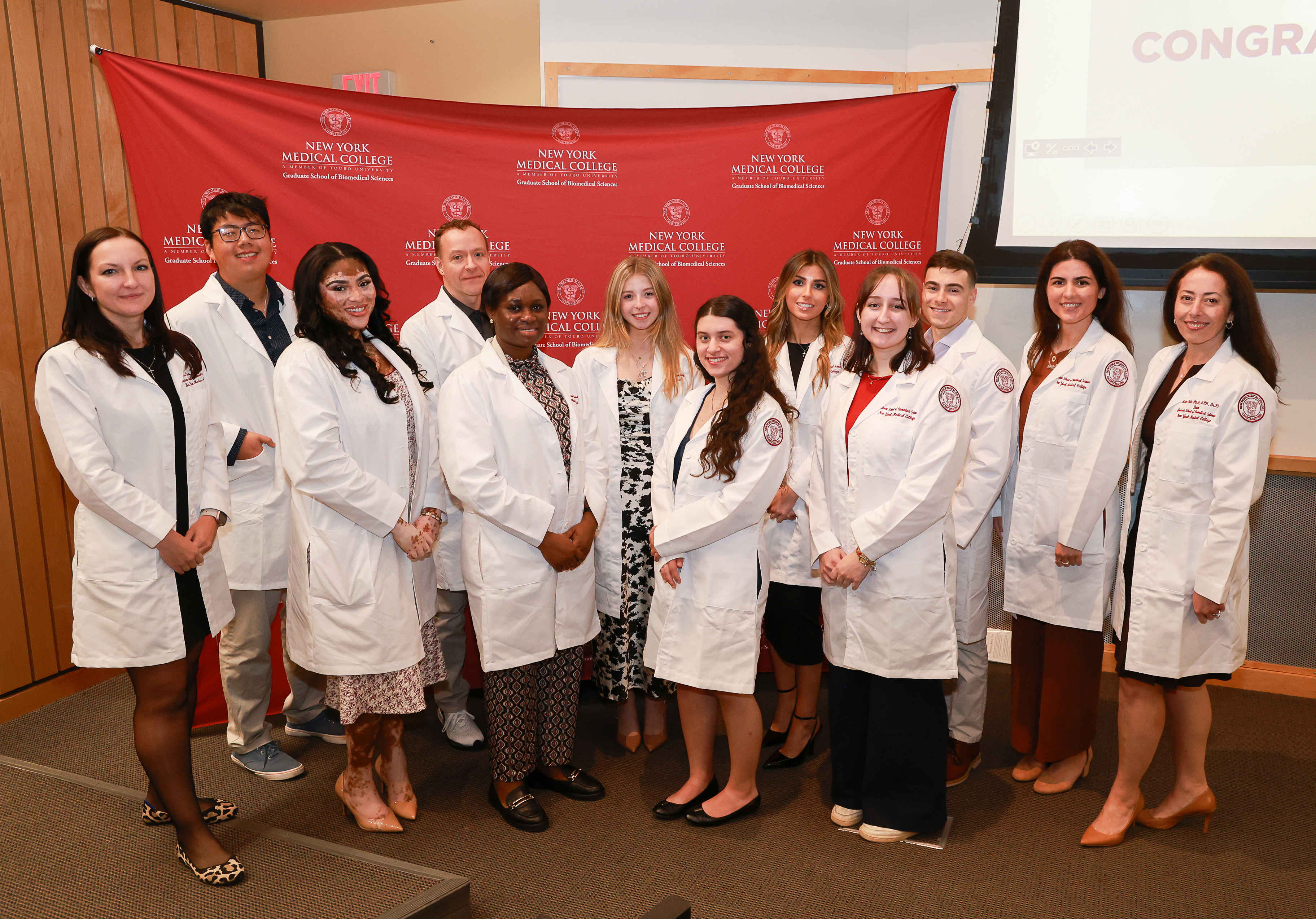 Students and faculty in while lab coats smiling in front of step and repeat.