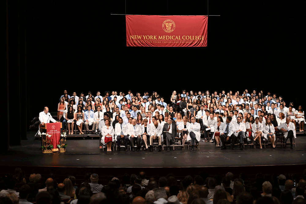 Man at podium in white coat on stage speaking to students in white coats