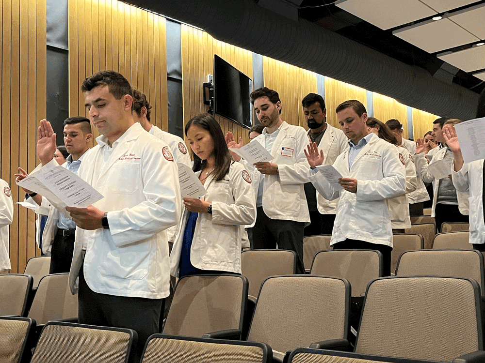 Medical students in white coat saying an oath as they lift their right hand and read from a folded paper in an auditorium.