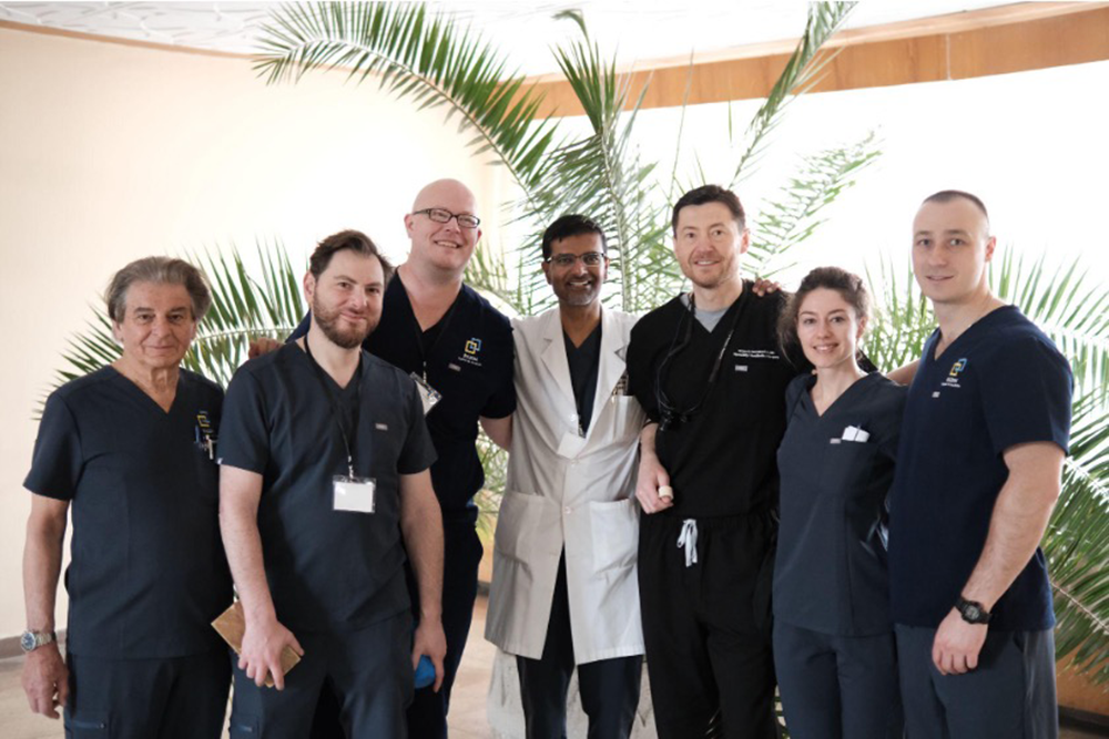 Medical professionals staring ahead smiling wearing scrubs in front of a palm tree indoor plant