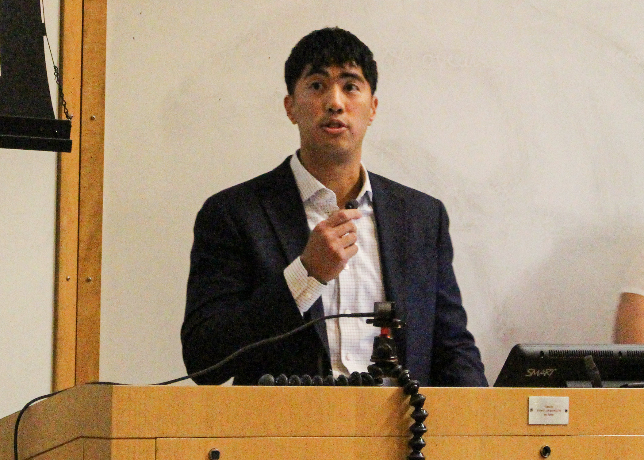 Male, standing at podium with microphone in hand, presenting to audience in auditorium