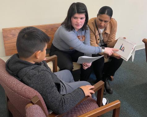 Two female speech-language pathology students, seated, showing a young boy, also seated, an illustration of a family as part of a pediatric language screening