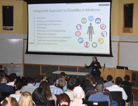 Professor standing in front of a classroom filled with students with a presentation slide on the board