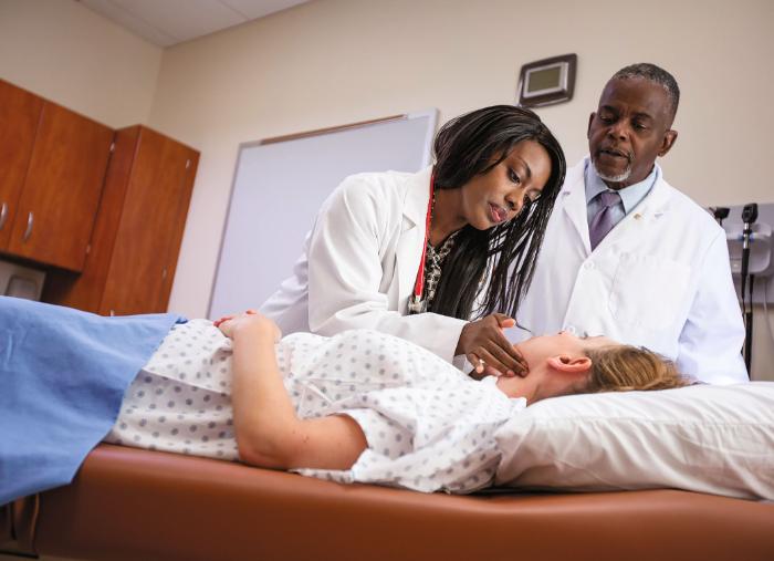A student examines a patient in a hospital bed with another doctor looking on.  